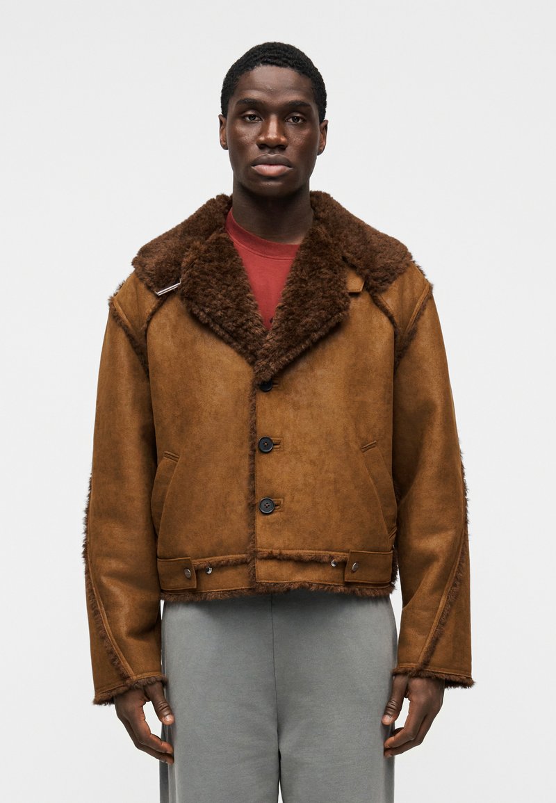 Καφέ δερμάτινο μπουφάν με μεγάλος γούνινος γιακάς, τρία μαύρα κουμπιά και πλαϊνές τσέπες. Σχεδιασμός με υφή, κοντός με ξέφτια στα άκρα.