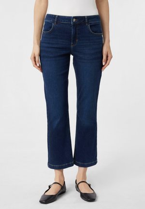 Vrouw draagt donkerblauwe rechte jeans en zwarte Mary Jane schoenen, staand tegen een effen lichte achtergrond.