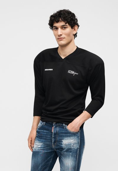 Νέος άνδρας με κατσαρά μαλλιά φοράει μαύρο μακρυμάνικο πουκάμισο με τα λογότυπα "DSQUARED2" και "ICONique" και φθαρμένα μπλε τζιν, με το χέρι στην τσέπη.