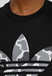 Černé bavlněné tričko s výrazným šedobílým maskovacím vzorem loga a třemi paralelními pruhy pod grafikou.