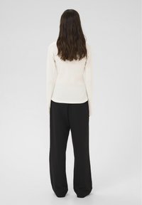 Kobieta z długimi brązowymi włosami, ubrana w białą bluzkę z długim rękawem i luźne czarne spodnie, stojąca plecami do obiektywu na jednolitym jasnym tle.