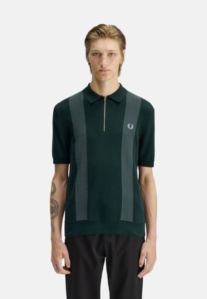 Joven con cabello claro que lleva una camiseta polo de manga corta verde oscuro con rayas verticales gris claro y un cuello con cremallera, de pie sin moverse.