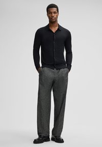 Schwarzes Langarmhemd mit Knopfleiste, kombiniert mit grauen strukturierten Hosen und schwarzen Schuhen. Das Outfit zeichnet sich durch ein körpernahes Design und eine maßgeschneiderte Silhouette aus.