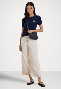 Moteris su ilgais tamsiais plaukais, vilkinti tamsiai mėlyną trumpomis rankovėmis megztinį su pynėmis, kreminės spalvos plataus kirpimo kelnes su mygtukais, tamsiai mėlyną per petį nešiojamą rankinę ir balerinas.