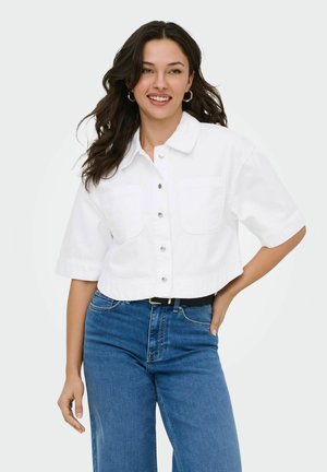 Frau mit dunklem Haar, die ein weißes, kurzärmeliges Hemd mit Knöpfen und blaue Jeans trägt, lächelt und steht mit einer Hand auf der Hüfte.