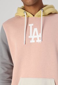 Sudadera con capucha que presenta una capucha amarilla en capas y un diseño en bloques de color: rosa, gris y crema. Cuenta con un logo blanco de "LA" en el pecho.