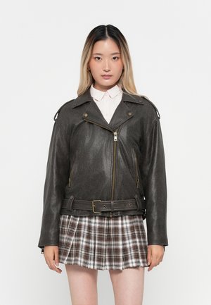 Bikerjack in donkergrijs leer met zilveren rits, zijzakken en een ceintuur op de taille. Draagt over een lichtgekleurde blouse en een geruite rok.