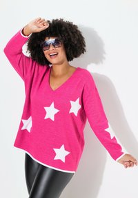 Rosa stickad tröja med vita stjärnmönster, V-ringning och lös passform. Matchas med svarta leggings med en kontrasterande vit fåll.