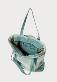 Zöld vászontáska tágas belső térrel, belső cipzáras zsebbel és a "HVISK" márkanevével a bélésre emboszolva.