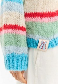 Sweter dziergany z poziomymi paskami w pastelowych kolorach: jasnoniebieskim, kremowym, różowym, zielonym i czerwonym. Metka zdobiona jest wzorem kokardy.