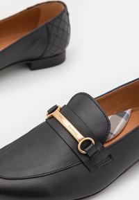 Mocassins en cuir noir avec détail boucle dorée marquée Barbour et semelle intérieure à carreaux, montrant les parties avant et arrière sur fond blanc.