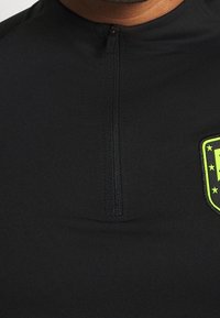 Schwarzes Sportshirt mit Viertelreißverschluss und einer strukturierten Applikation mit grünem Umriss und Sternen. Hergestellt aus glattem, dehnbarem Stoff.