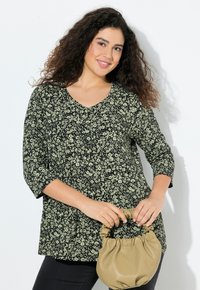 Schwarze Blumenshirt mit Dreiviertelärmeln, V-Ausschnitt und grünen Mustern. Das Model hält eine kleine beige Handtasche.