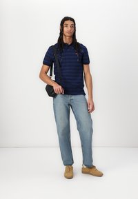 Polo shirt cu dungi bleumarin, blugi deschiși la culoare și pantofi sport bej. Accesoriile includ o geantă mică crossbody neagră.