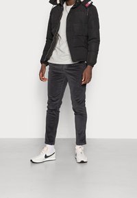 Giacca a vento nera con zip e cappuccio, maglietta grigia a girocollo, pantaloni in velluto a coste grigio scuro e sneakers bianche Nike con dettagli neri.