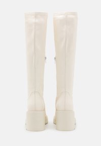 Krémové boty až po kolena vyrobené z hladkého materiálu, se čtveratým podpatkem, bočními zipy a viditelnými švy.