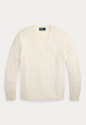 Jeune homme portant un pull en maille torsadée crème sur une chemise bleue, un pantalon en velours côtelé marron foncé, des chaussettes grises et des chaussures habillées marron, debout les mains dans les poches.