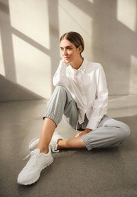 Camicia bianca con colletto borchiato, pantaloni jogger grigi chiari e sneakers bianche. Posizione seduta, pavimento in cemento liscio, ombre morbide.