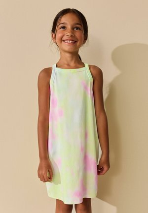 Smiling young girl with dark hair wearing a sleeveless pastel tie-dye dress against a beige background.