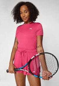 Top de desporto rosa com mangas curtas e um logótipo branco, combinado com calções rosa iguais com cordão. Racket de ténis preto segurado na mão.