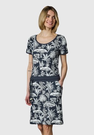 Frau trägt ein schwarzes Kleid mit weißen tropischen Pflanzenmustern, kurzen Ärmeln, rundem Ausschnitt und einem Taillengürtel mit Quasten.