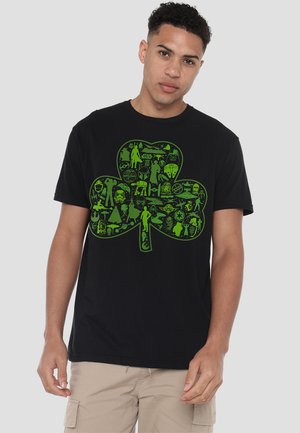Junger Mann, der ein schwarzes T-Shirt mit einem großen grünen Kleeblatt aus Silhouetten von Star Wars-Charakteren trägt, beigen Cargo-Shorts und vor einem grauen Hintergrund steht.
