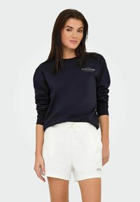 Mørkeblå sweatshirt med rund hals og hvid grafik, parret med hvide shorts med snor. Glat stof, afslappet pasform, casual stil.
