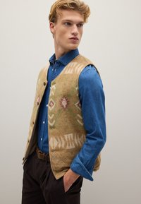 Beige gemusterte Weste mit geometrischen Designs, über einem blauen Jeanshemd getragen. Die Weste hat eine weiche Textur und einen Knopfverschluss.