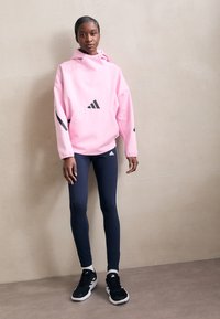 Rožinė kapučino džemperis su juodais akcentais, derinamas su tamsiai mėlynomis leginso ir juodais sportiniais batais, visi daiktai su Adidas logotipu.