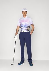 Pánske golfové oblečenie pozostávajúce z trička s krátkym rukávom v ružovej a modrej farbe s grafikou, námorníckych nohavíc, bieleho opasku, bielej čiapky a golfovej palice.