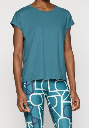 Person iført en teal kortærmet top og teal leggings med store abstrakte hvide og mørkeblå geometriske bogstaver.