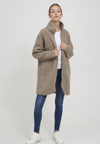 Beige fleecjas met hoge kraag, open voorkant en een relaxed fit, gecombineerd met donkerblauwe jeans en witte sneakers. Gladde textuur.