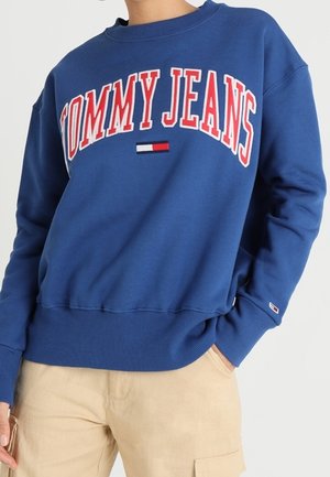 Sweat-shirt bleu avec "TOMMY JEANS" en lettres rouges et blanches et un petit logo de drapeau, associé à un pantalon beige.