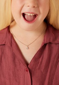 Zilveren ketting met een hartvormige hanger, versierd met kleine heldere steentjes. Draagt over een textuurrijke bordeauxrode blouse met een kraag.