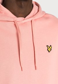 Uomo che indossa un felpa leggera di colore rosa chiaro con cappuccio e un piccolo logo di un uccellino giallo sul lato sinistro del petto, su uno sfondo semplice.
