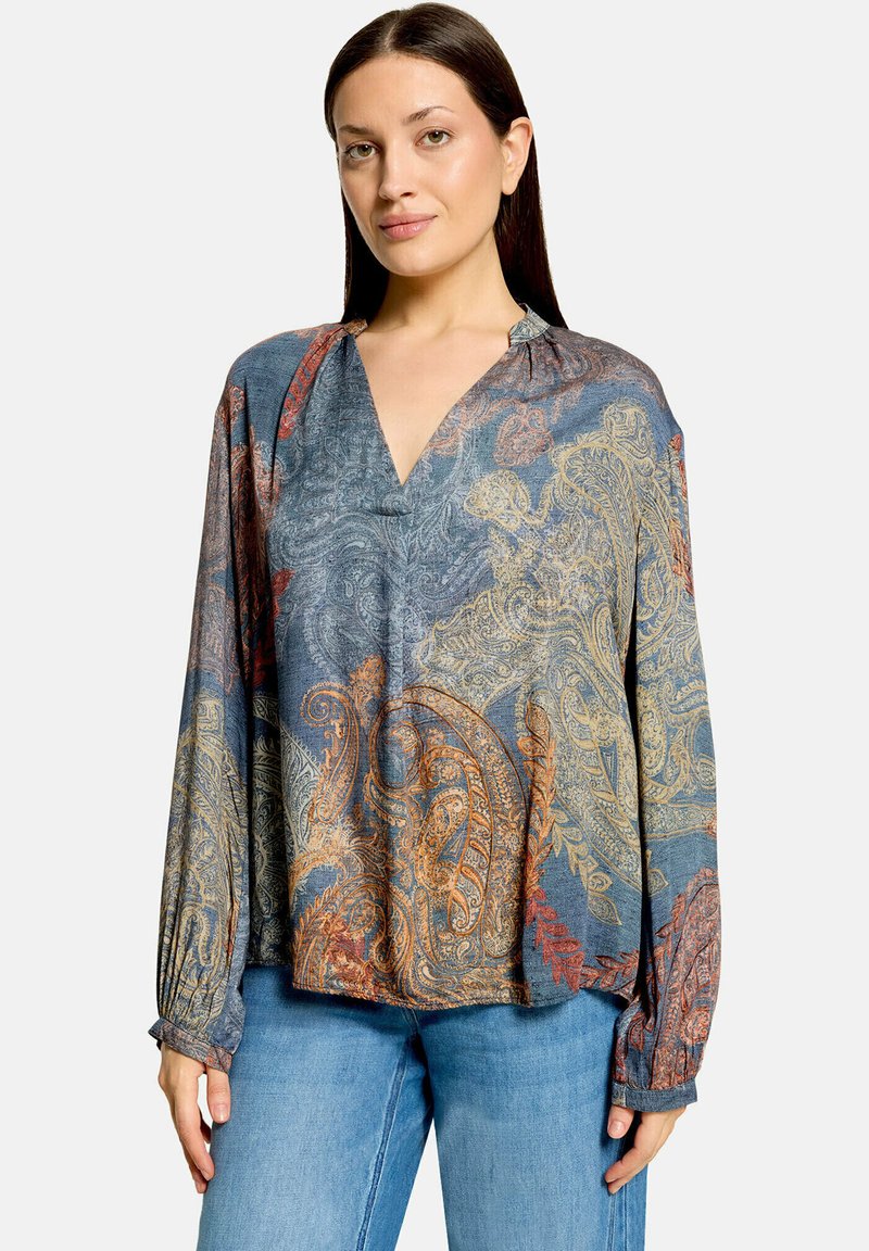 Femme aux longs cheveux foncés portant un chemisier bleu à motif cachemire à manches longues et un jean bleu sur un fond uni.