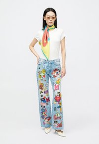 Jeans boca de sino em denim azul claro com remendos coloridos; combinados com uma blusa branca ajustada e um lenço de seda multicolorido. Saltos brancos.