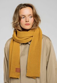 Musztardowo-żółty, dziany szalik o teksturowanym wykończeniu z brązową skórzaną metką, swobodnie zawieszony na szyi, na tle w neutralnych kolorach.