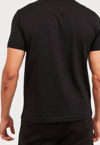 Hombre vestido con una camiseta negra de manga corta lisa, visto desde atrás sobre un fondo claro y liso.