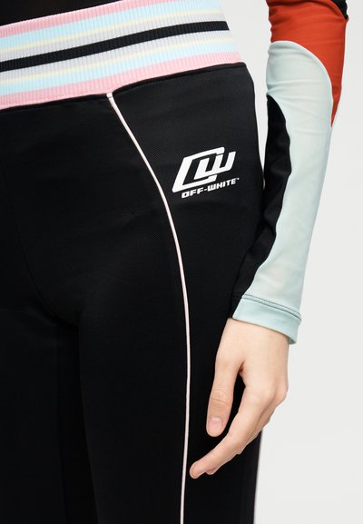 Κοντινό πλάνο ατόμου που φορά μαύρο παντελόνι Off-White με λευκό λογότυπο και λεπτή λευκή γραμμή, καθώς και πολύχρωμο ριγωτό τοπ με μανίκι σε χρώματα μπλοκ.