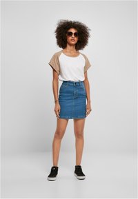 Hvit og beige raglanermet skjorte, blå denim miniskjørt, svarte joggesko, store brune solbriller; modellen står mot en lys grå bakgrunn.