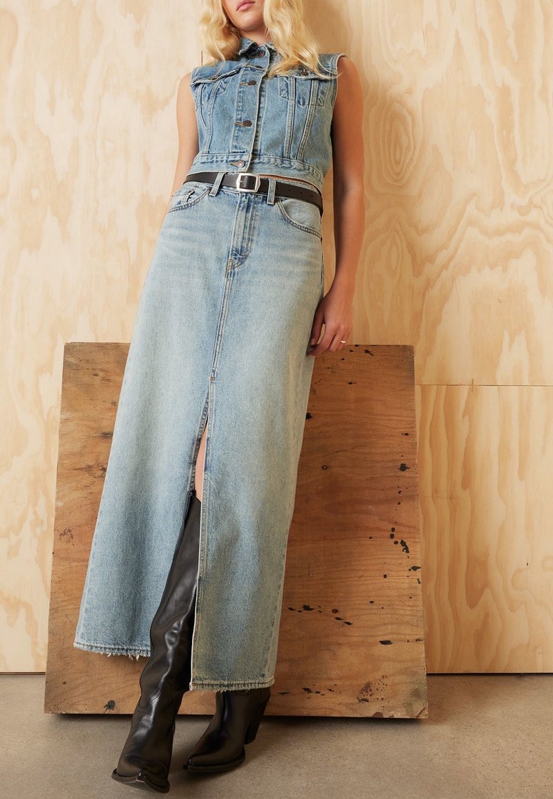 Pitkä denimgdressi, jossa on olkaimeton muotoilu, vaaleansininen väri, edessä halkio ja musta vyö. Yhdistettynä korkeisiin mustiin korkokenkiin.