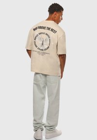 Beige oversized t-shirt med grafisk tekst og due-design på ryggen. Sættes sammen med lysegrønne bukser og hvide sneakers. Simpel, afslappet stil.