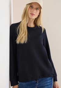 Marineblauwe sweatshirt met lange mouwen, een gestructureerde brei, ronde halslijn en details met zijkoord. Gecombineerd met blauwe jeans.