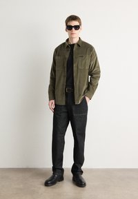 Olive green button-up shirt with two chest pockets, black t-shirt underneath, dark denim trousers, and black shoes. Sunglasses complete the look.