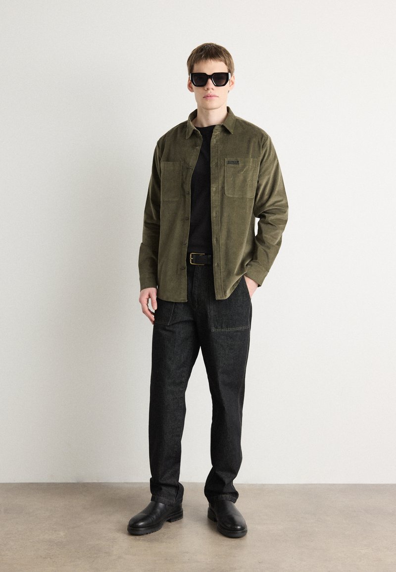 Olijfgroene knopshirt met twee borstzakken, zwart T-shirt eronder, donkerdenim broek en zwarte schoenen. Zonnebril maakt de look compleet.