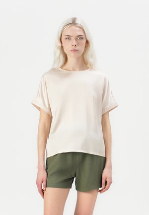 Lys beige kortermet topp med løs passform, rund hals og rullende ermer, kombinert med mørk grønn shorts. Silkeaktig tekstur og minimalistisk design.