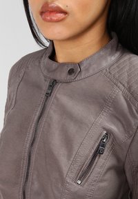 Chaqueta de cuero sintético gris con un cuello alto, detalles de costura visibles y dos bolsillos con cremallera en la parte frontal. Textura suave.