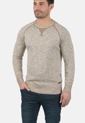 Lys brun strikkegenser med et teksturert mønster, ribbede mansjetter og kant. Har en rund hals og kontrasterende beige kant.
