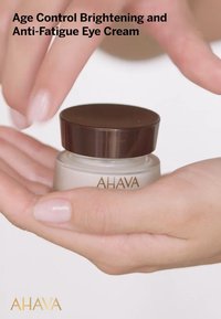 Crema per gli occhi AHAVA Age Control in un vaso di vetro trasparente con coperchio marrone, tenuta in mano. Texture liscia, di colore crema chiaro.